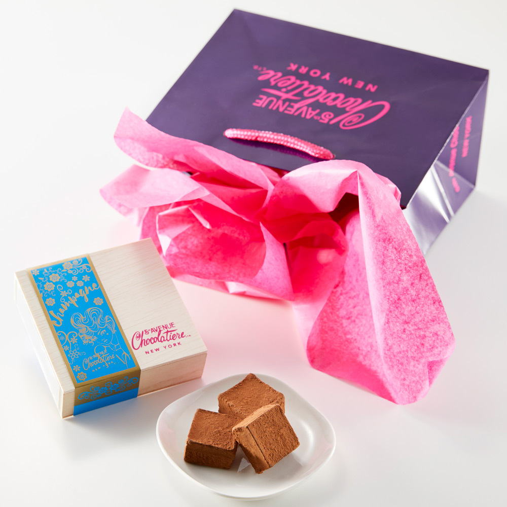 Box of chocolates beside a plate with chocolate pieces, next to a purple gift bag with pink tissue paper.