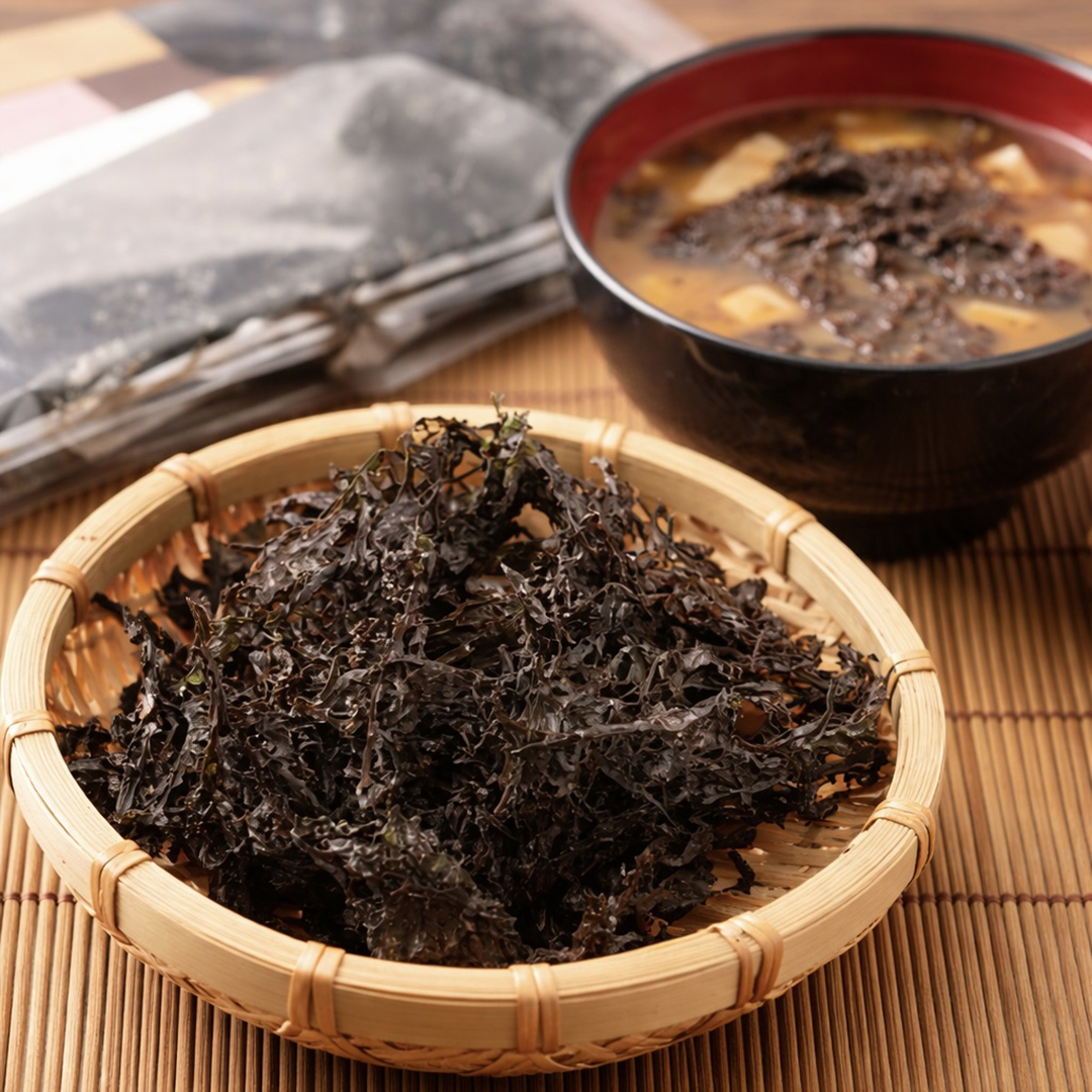 Dried seaweed in a bamboo basket next to a bowl of soup containing seaweed on a bamboo mat.