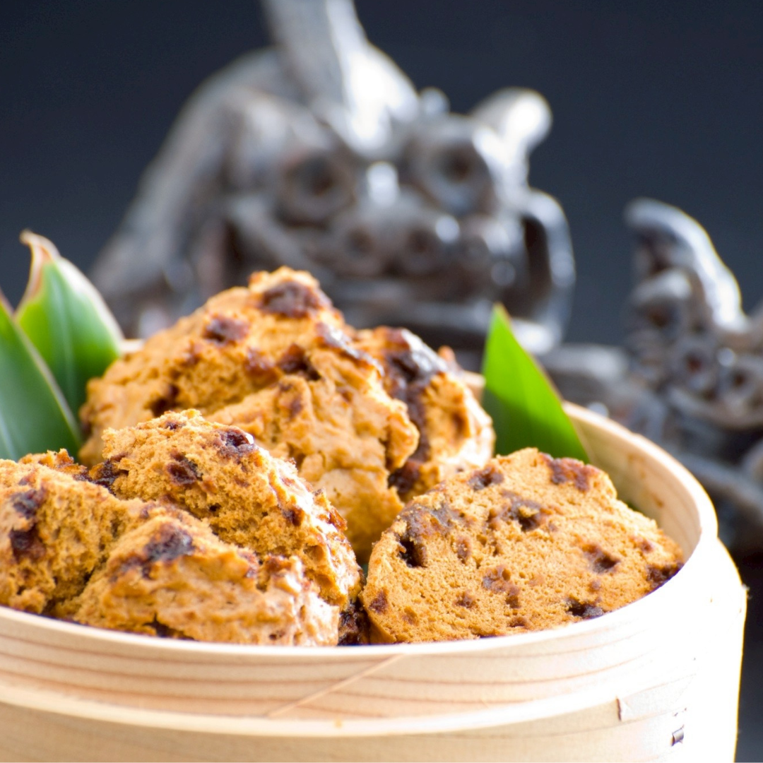 A bowl of brown cookies with chocolate chunks, garnished with green leaves, set against a dark background with a blurred sculpture.