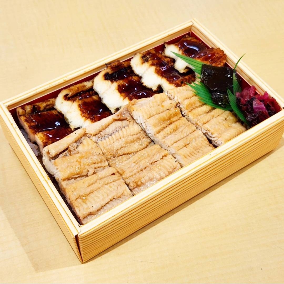 A wooden box with sliced grilled eel, rice, and garnishes, including a green leaf and pickled vegetables, arranged neatly inside.