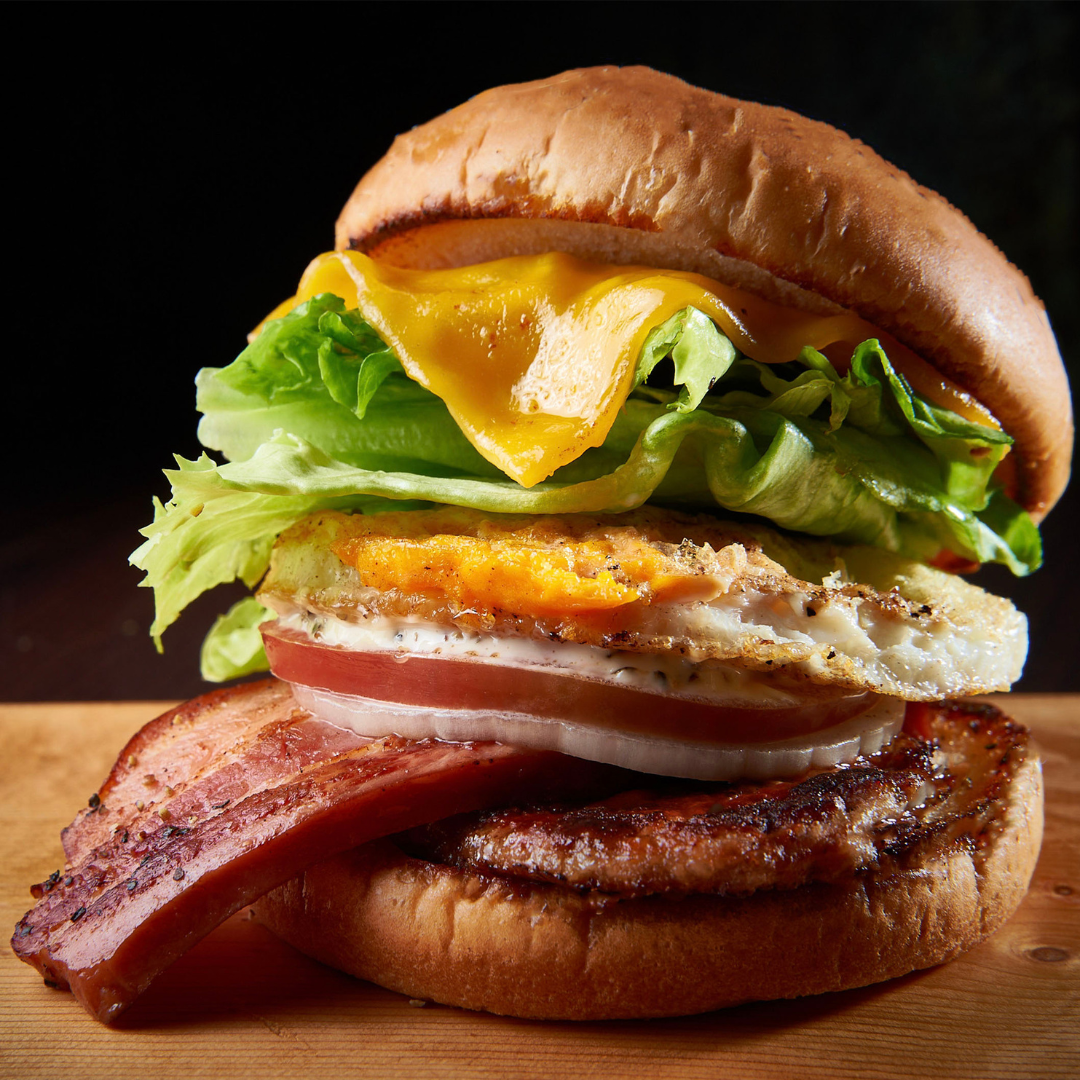 A loaded burger with lettuce, cheese, egg, tomato, onion, bacon, and a patty on a wooden board.