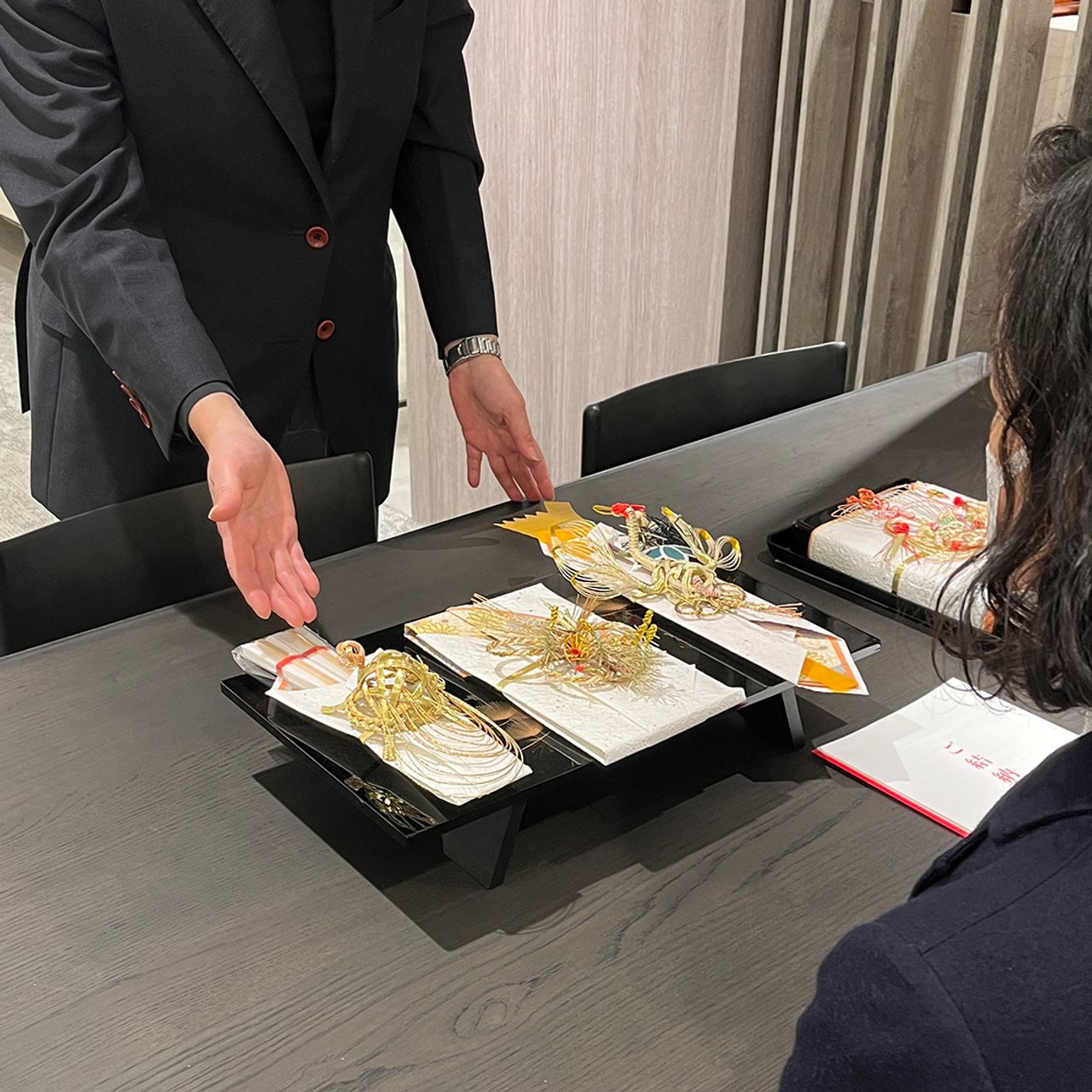 テーブルに着席したお客様へ、儀礼用の華やかな祝儀袋を載せた盆を差し出す様子。格式ある場面を想起させます。