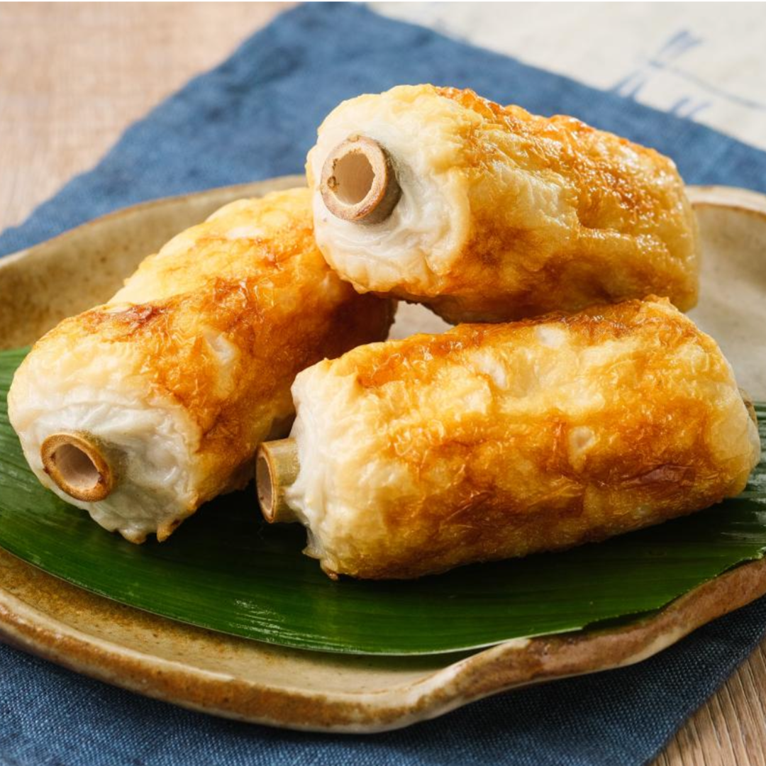 Three grilled chikuwa fish cakes on a plate with a green leaf base. They are golden brown and cylindrical, placed on a blue cloth.