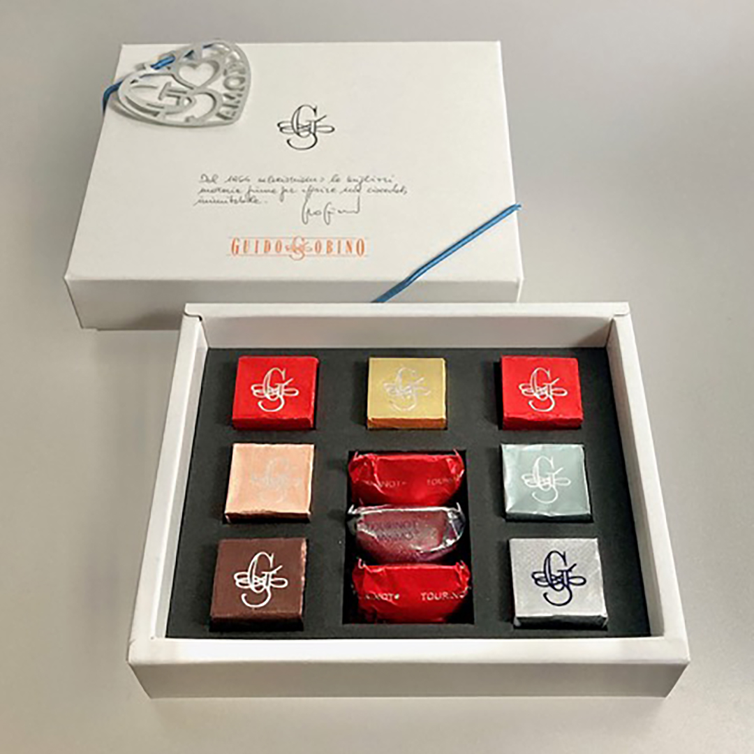 Open box of assorted chocolates with colorful wrappers, arranged in a 3x3 grid. White box features a decorative silver heart charm.