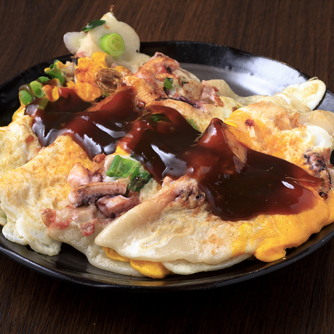 A plate of takoyaki omelette topped with a rich brown sauce and garnished with green onions, served on a dark wooden table.