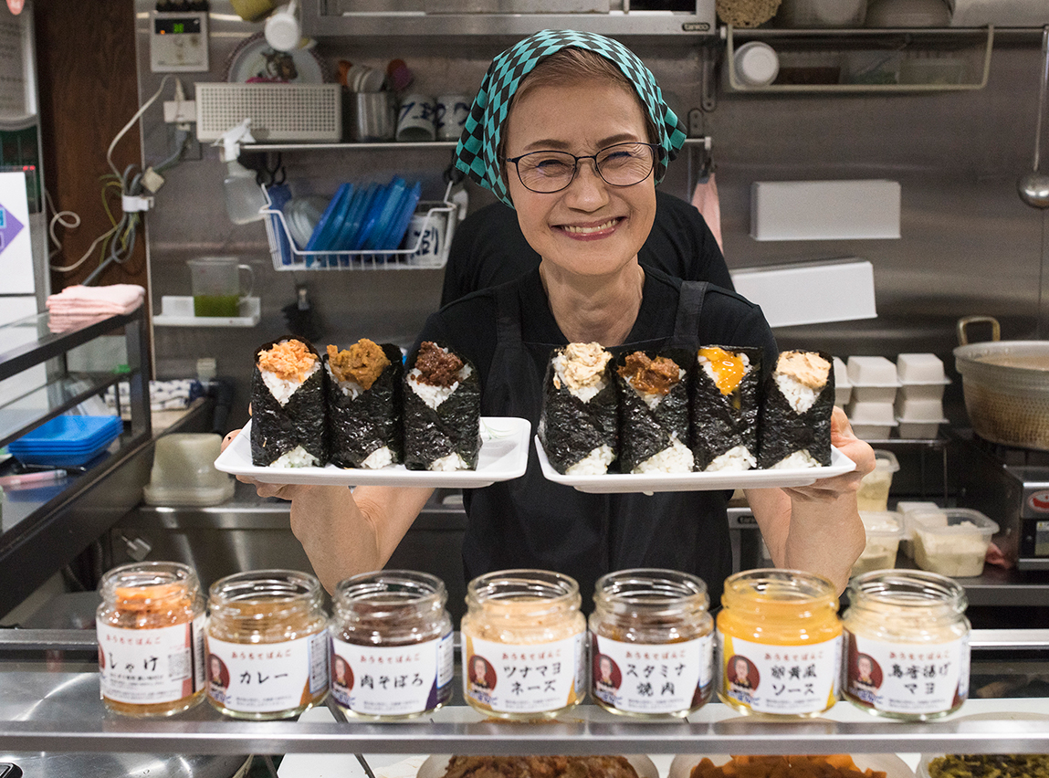 Smiling person holding plates of onigiri in a kitchen, with jars of various toppings displayed in front.