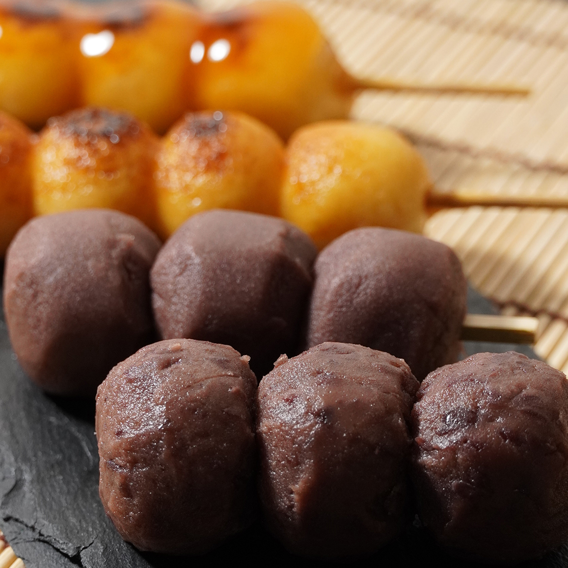 Close-up of skewered Japanese sweets, including brown bean paste balls in the foreground and glazed dango in the background on a slate plate.