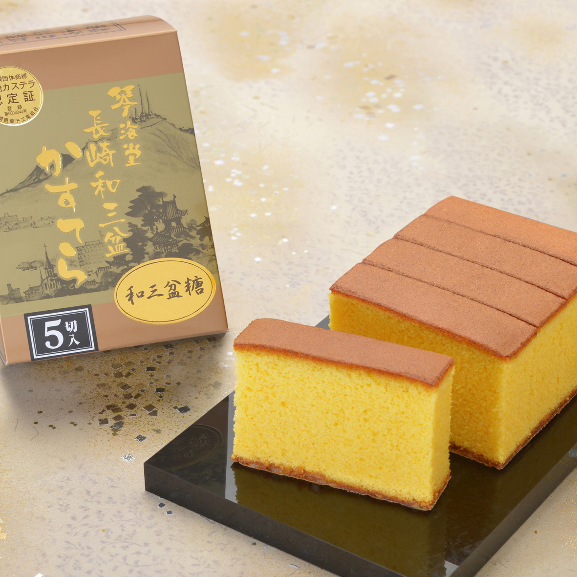 Golden castella slices on a black plate next to a decorative box with Japanese text, set on a speckled beige surface.