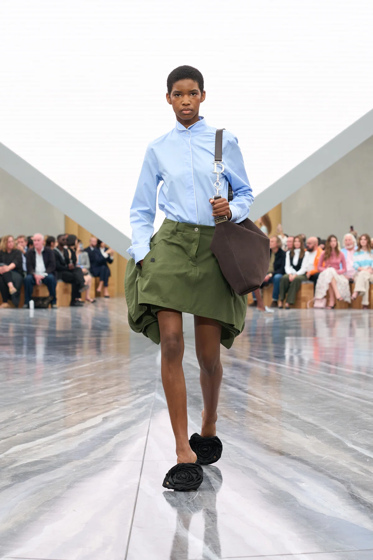 Model in light blue shirt, green skirt, and black shoes walks on a reflective runway, holding a brown bag, with audience seated in background.