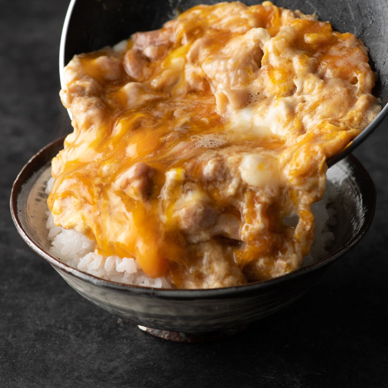 A bowl of rice topped with creamy, golden egg and chicken mixture, served on a dark surface.