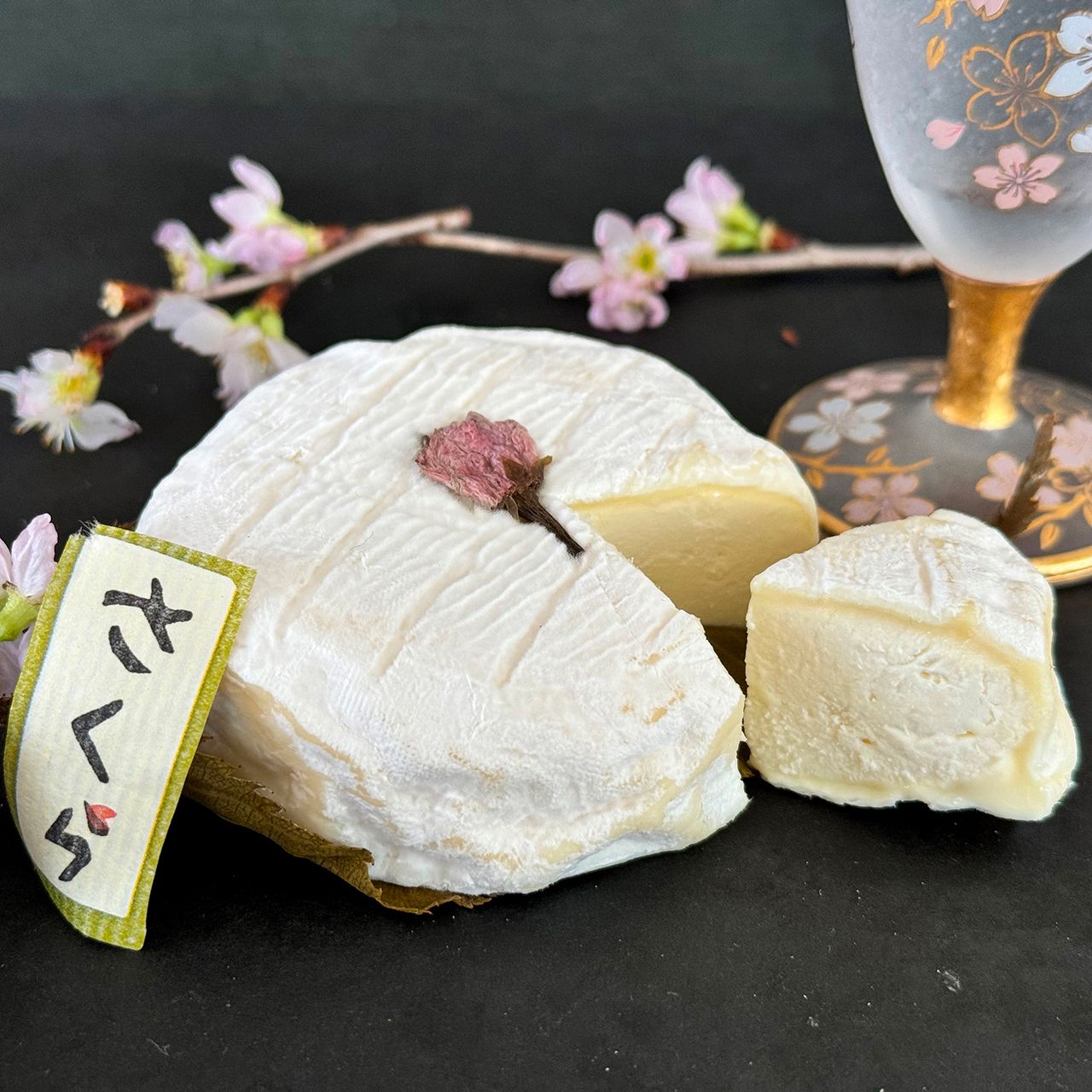 Round white cheese with a cherry blossom on top, a cut slice next to it, decorated glass, cherry blossoms, and a "さくら" label.