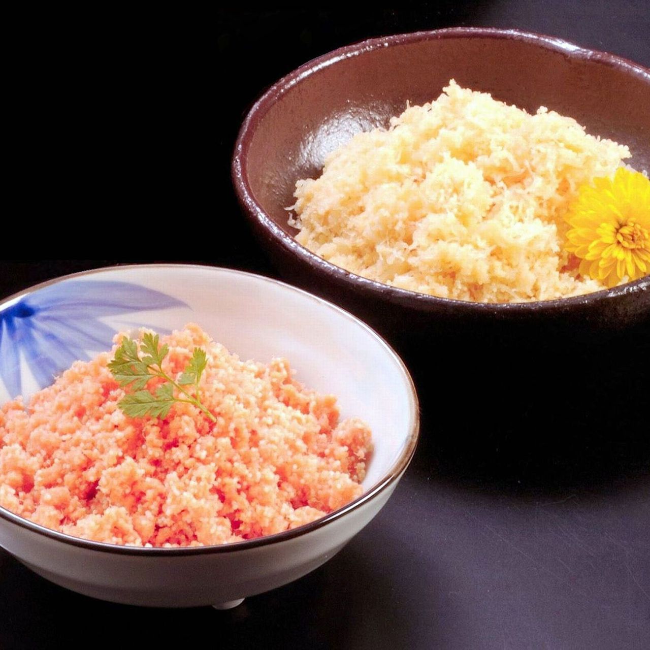 Two bowls of grated food: one with orange and another with pale yellow shreds. A yellow flower adorns the latter. Black background.