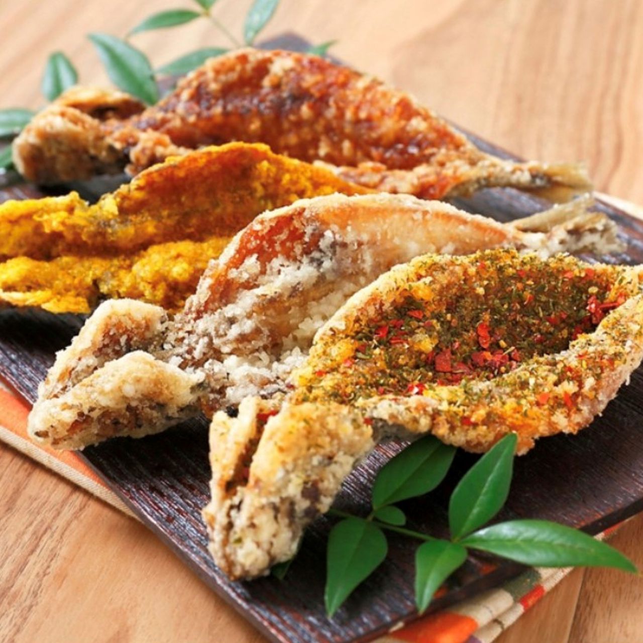 A platter of various seasoned and fried fish pieces on a dark rectangular plate, garnished with small green leaves.