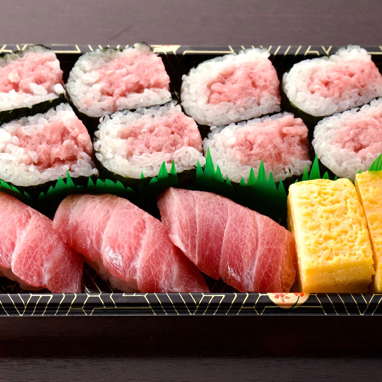 A sushi tray with tuna rolls, tuna nigiri, and tamago slices on a dark surface.