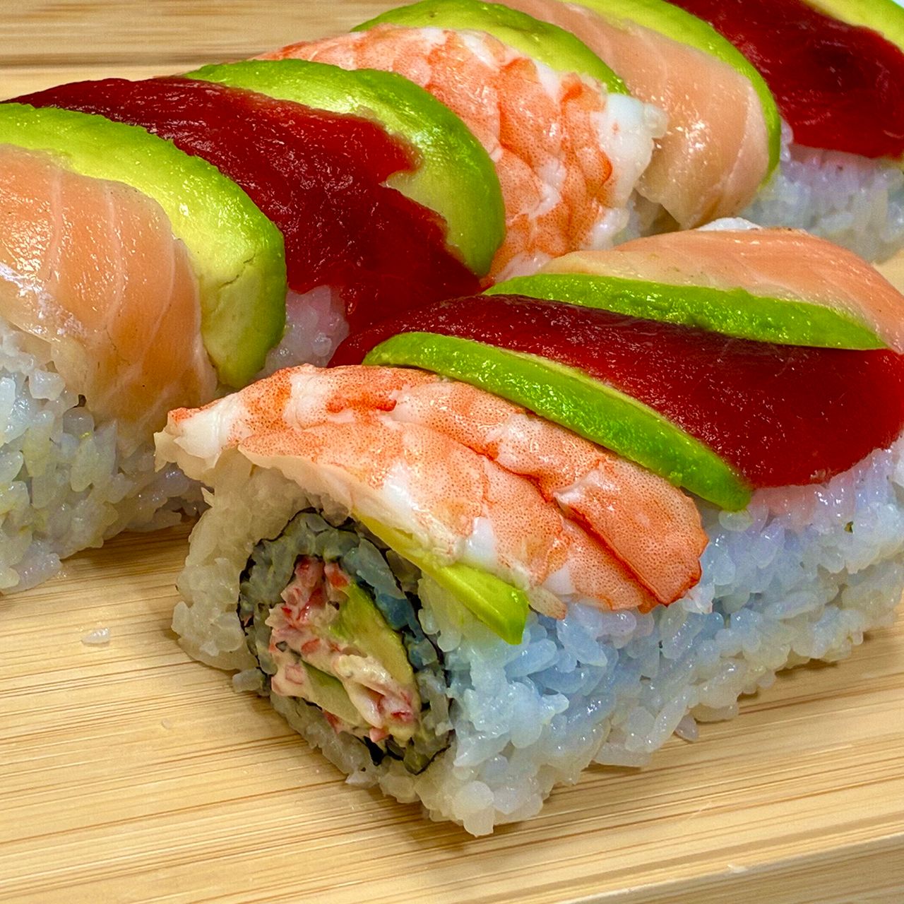 Close-up of sushi rolls topped with avocado, tuna, shrimp, and salmon on a wooden board. Rolls filled with crab, cucumber, and avocado.