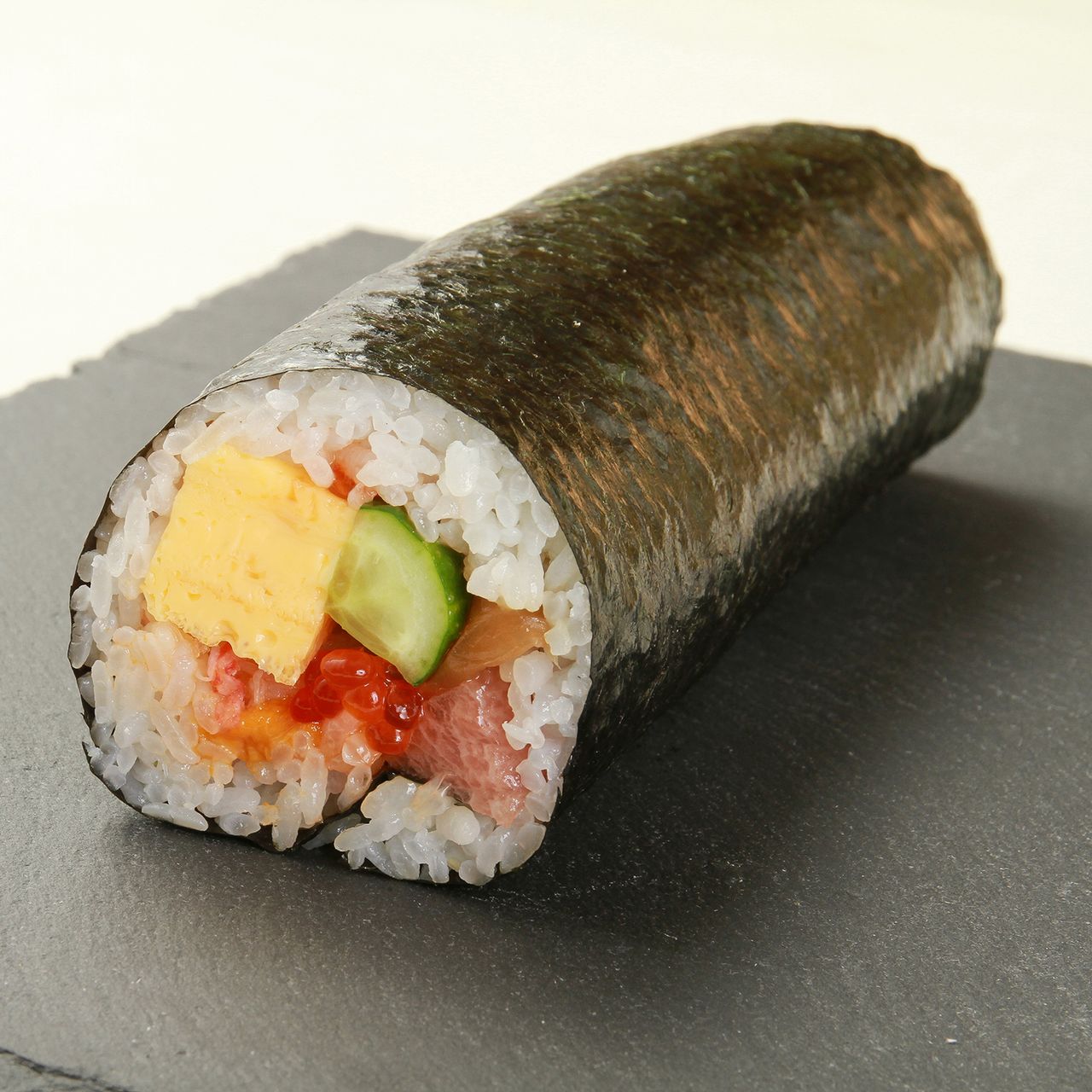 海苔で巻かれた寿司ロール。白いご飯に、きゅうり、玉子、魚、赤い魚卵を合わせ、ダークカラーのスレート皿に盛り付けられています。