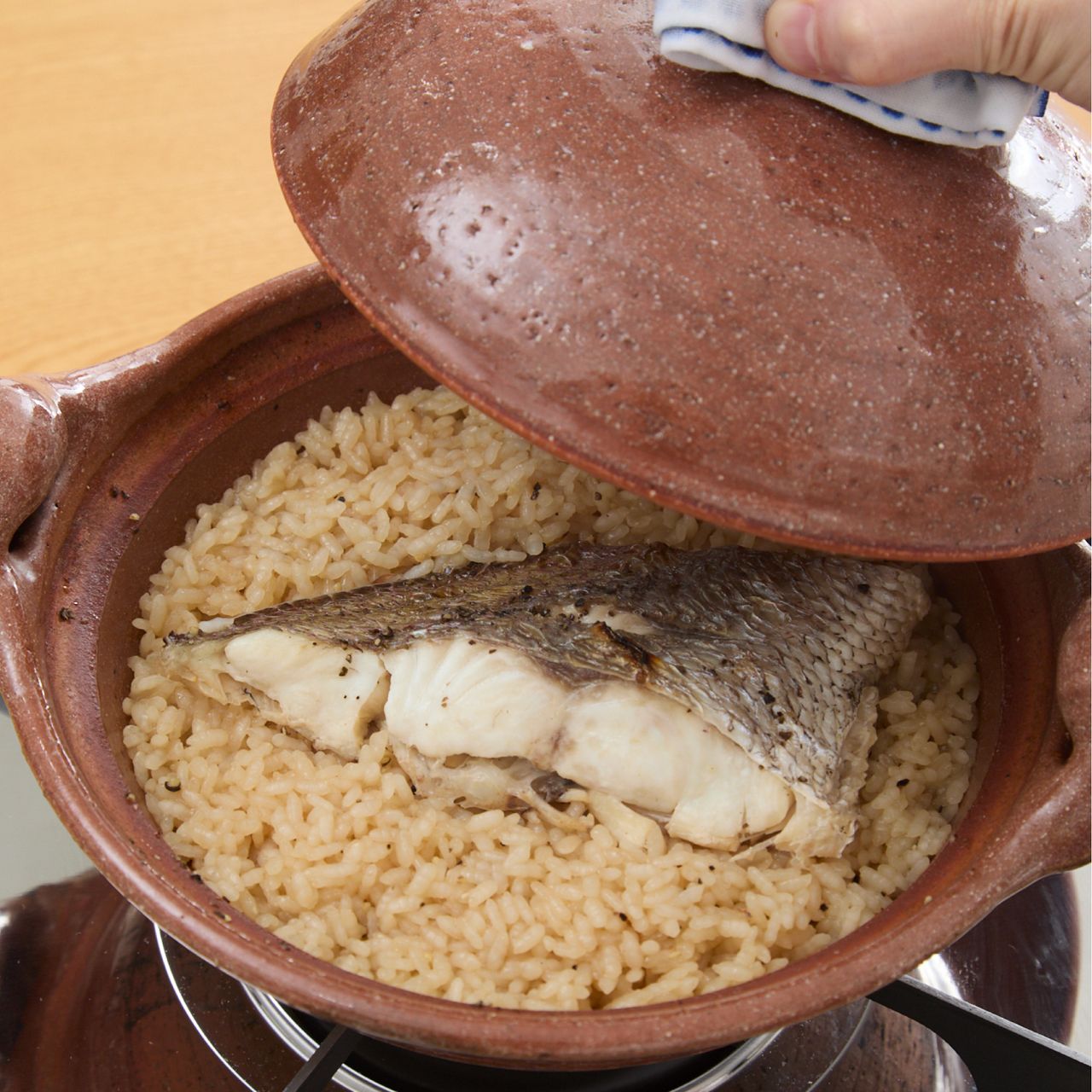 A hand lifts the lid of a clay pot, revealing a cooked fish fillet on a bed of seasoned rice.