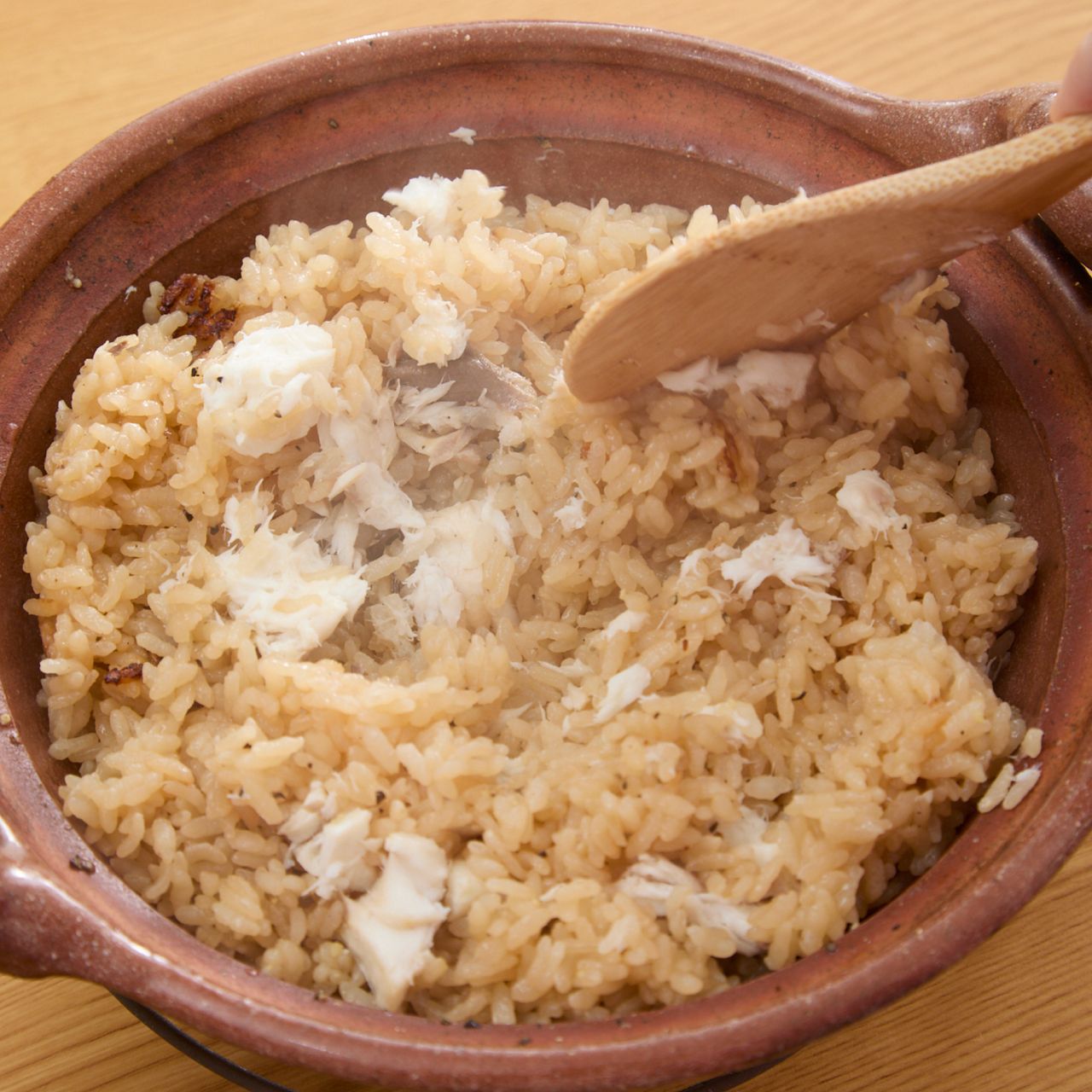 Clay pot filled with steamed rice being stirred with a wooden spoon, with visible steam rising.