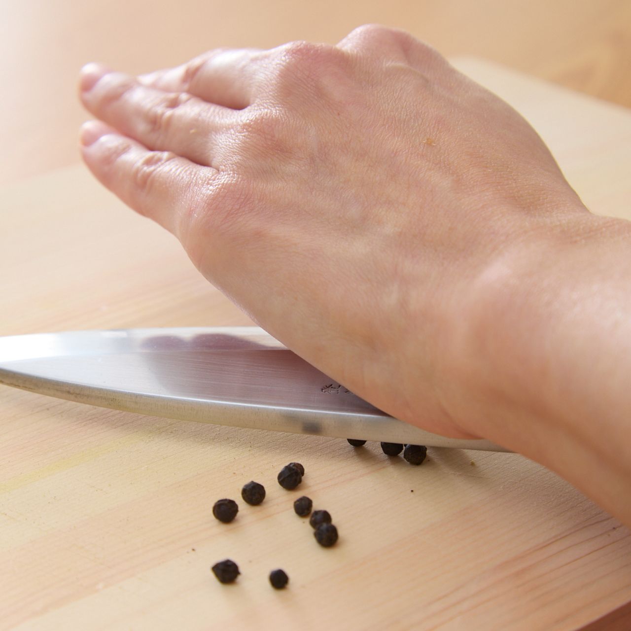 Hand pressing a knife blade on black peppercorns on a wooden cutting board.