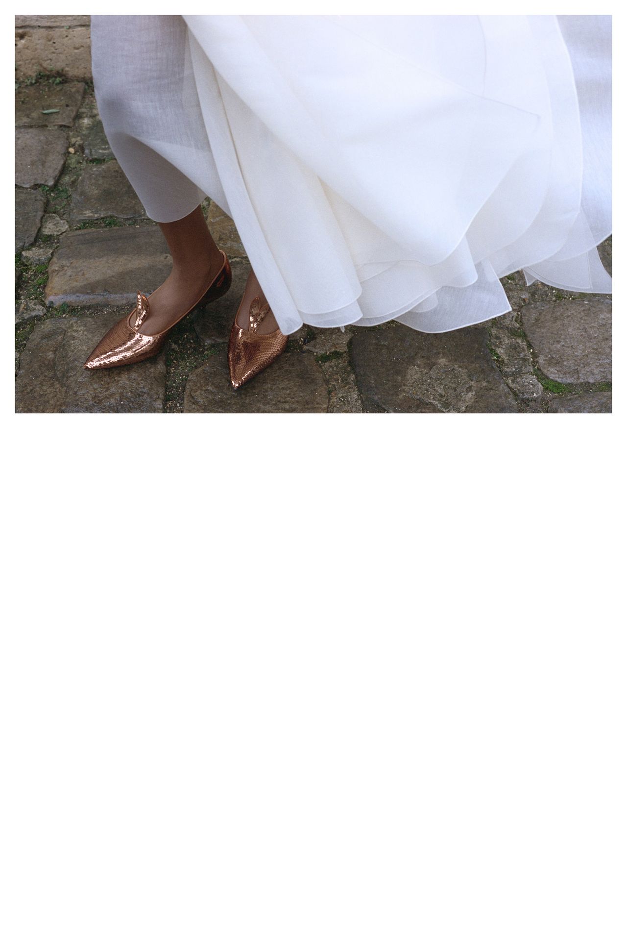Person in a flowing white dress, stepping on a cobblestone path, wearing shiny bronze loafers adorned with a small chain detail.