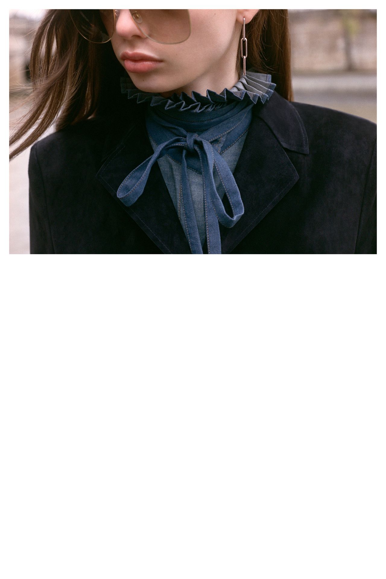 Woman wearing sunglasses, dark coat, and blue collared shirt with a ruffled neckline and bowtie detail.