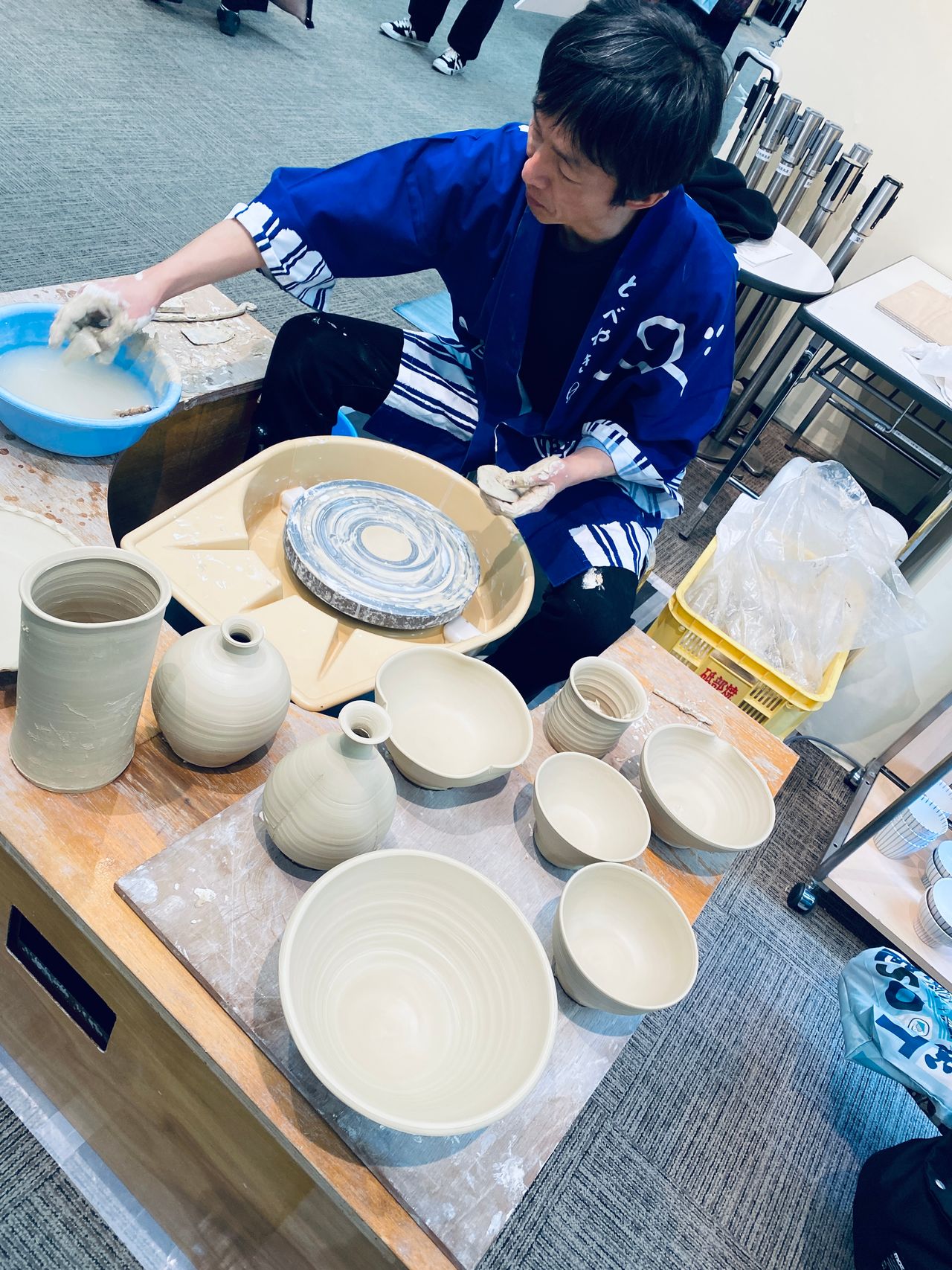 Person in a blue robe shaping pottery on a wheel, surrounded by finished beige ceramic bowls and vases on a table.