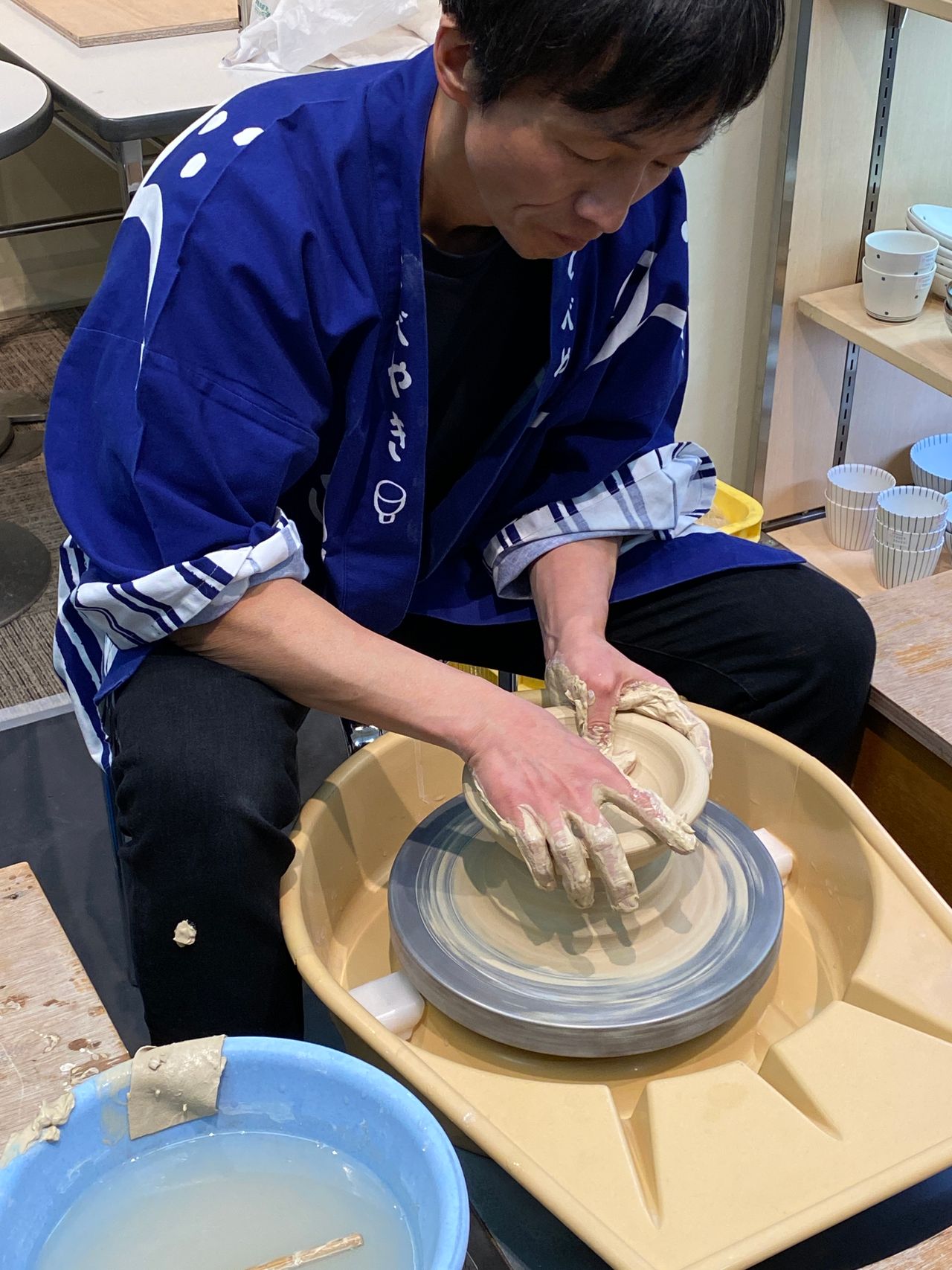 Person in a blue kimono shapes clay on a pottery wheel, with water and tools nearby, focused on creating a ceramic piece.