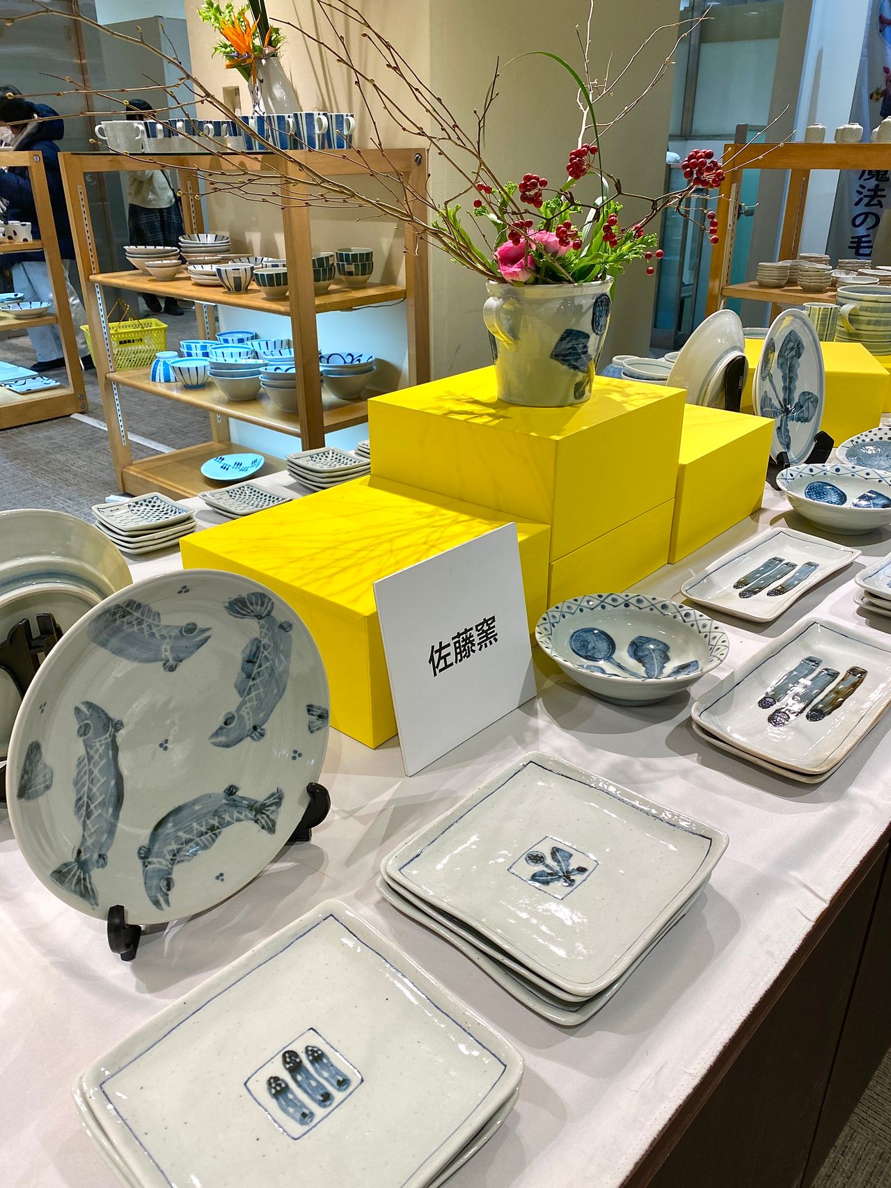 Display of ceramic plates and bowls with blue fish patterns on a white table, accented by yellow stands and floral arrangements.