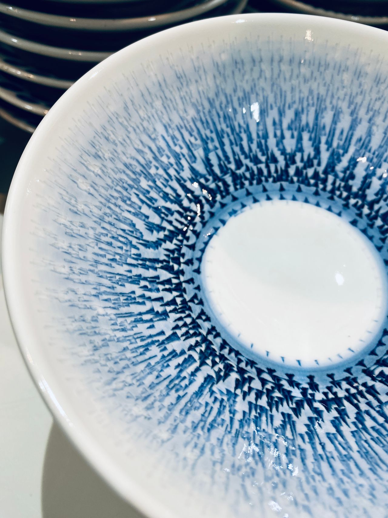 Close-up of a white bowl with intricate blue radial patterns, resembling abstract art. Stacked plates are visible in the background.
