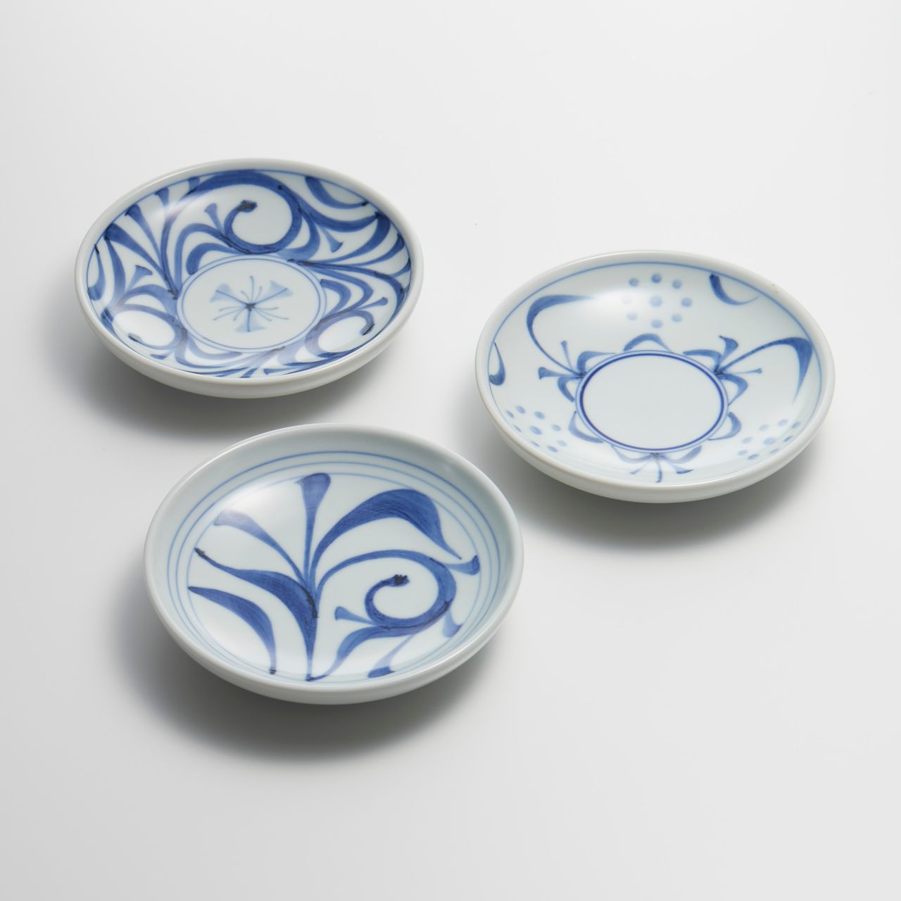 白地に青の花柄や抽象模様が描かれた、小さな陶器の皿3枚が、白い面の上に三角形に配置されています。