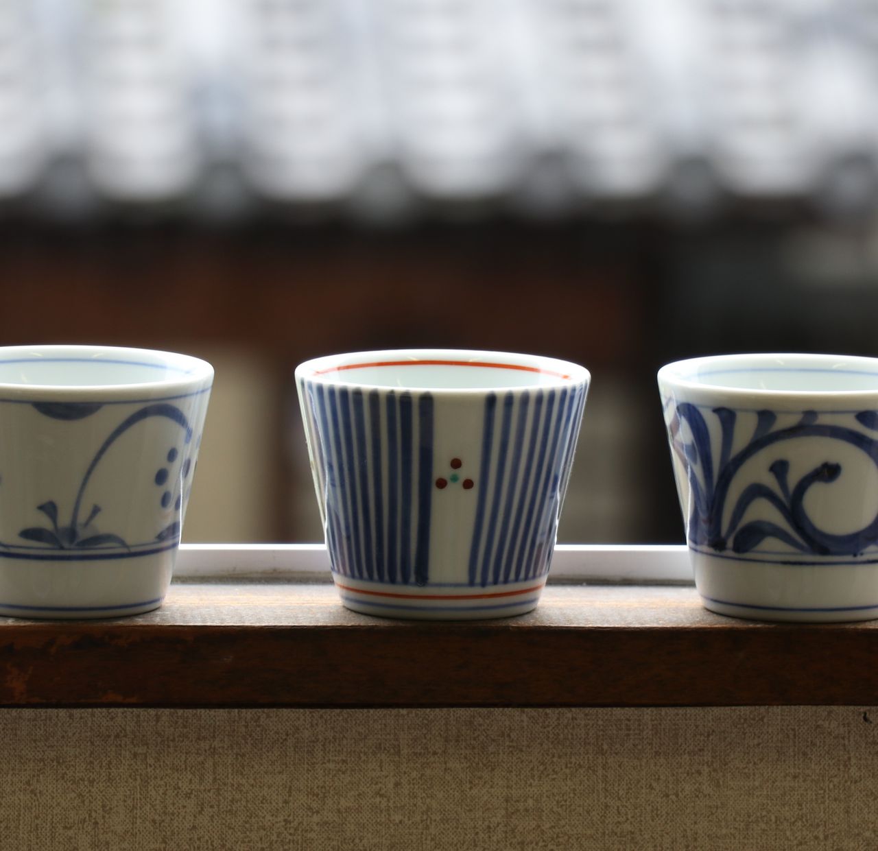 Three ceramic cups with blue and white patterns sit on a windowsill, with a blurred background of a traditional building.