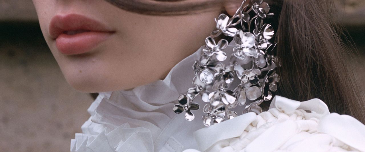 Close-up of a person wearing a ruffled white blouse and large metallic floral earrings, with long hair partially covering their face.