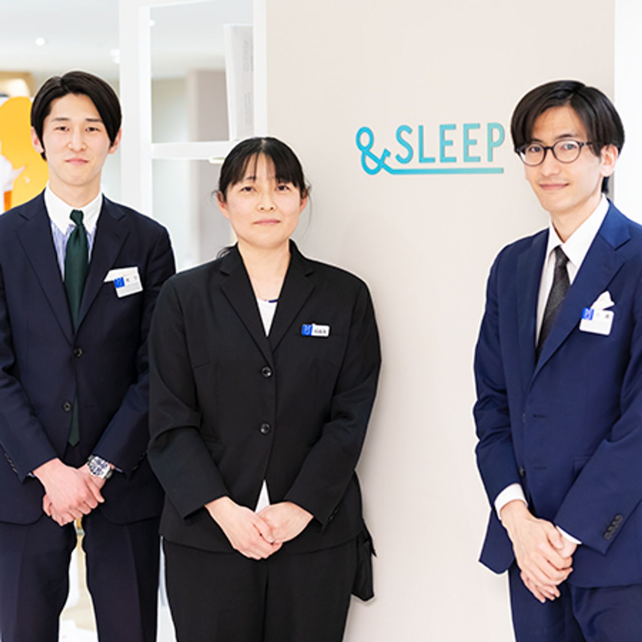 Three people in formal attire stand by a wall with the word "&SLEEP" on it. They are wearing name tags and smiling.