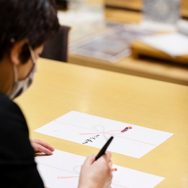 A staff member wearing a mask carefully handwrites a formal message on decorative paper, showcasing expert calligraphy.