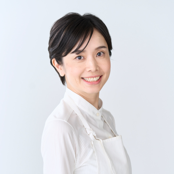 Smiling person with short dark hair wearing a white shirt and apron, standing against a light background.