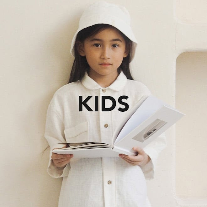 Child in white outfit and hat holding an open book, standing against a light background with the word "KIDS" overlaid.